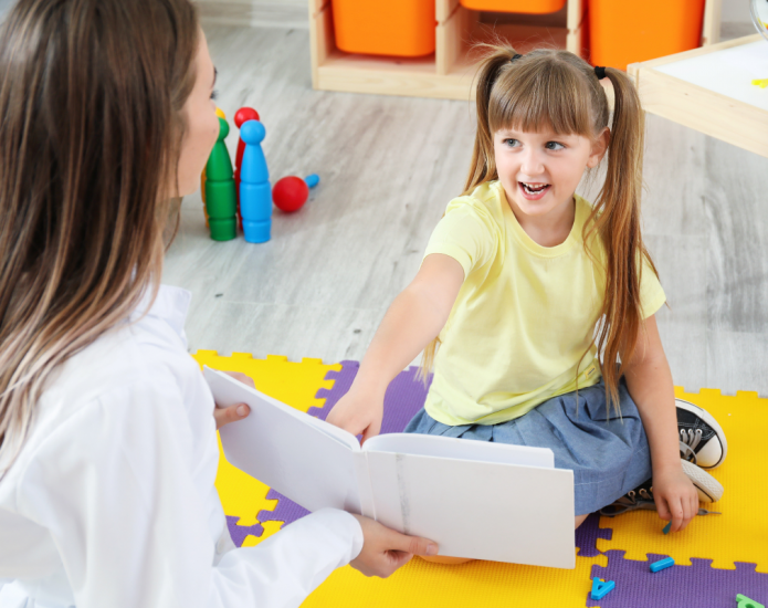 Young girl pointing to and reading from book held by speech therapist