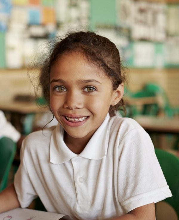 Young smiling girl in white school uniform sitting at desk in classroom