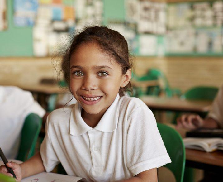 Young smiling girl in white school uniform sitting at desk in classroom