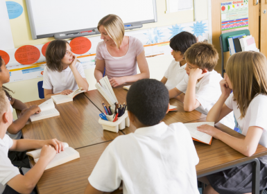 Teacher sitting at desk speaking with group pf primary school students