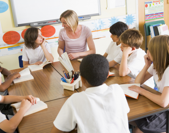 Teacher sitting at desk speaking with group pf primary school students