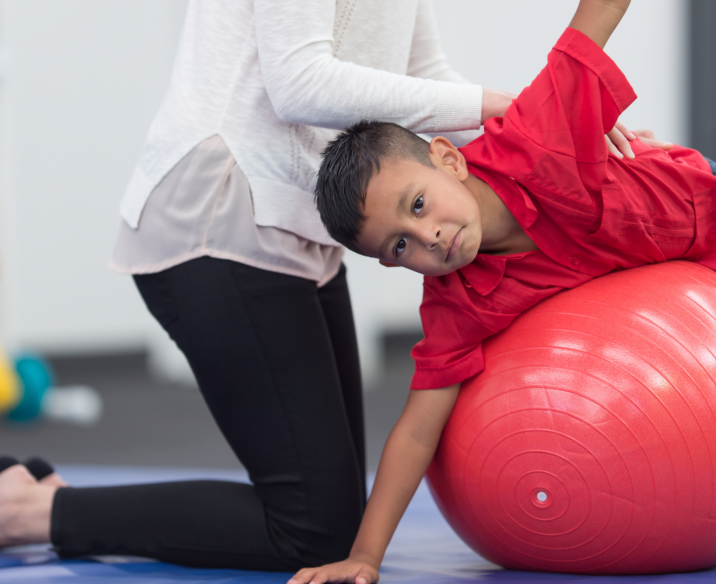 Therapist helping schoolboy balance on stomach on top of exercise ball