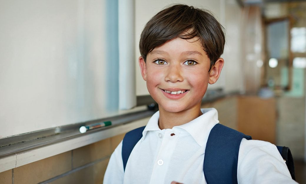 Young boy with brown eyes smiling next to whiteboard at school