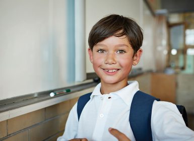 Young boy with brown eyes smiling next to whiteboard at school