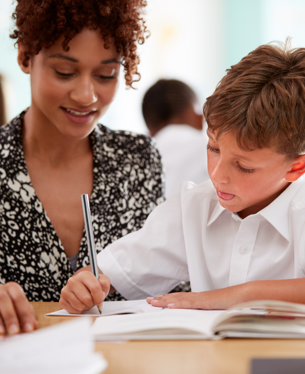 Teacher sits next to student with tongue poking out while concentrating on writing task