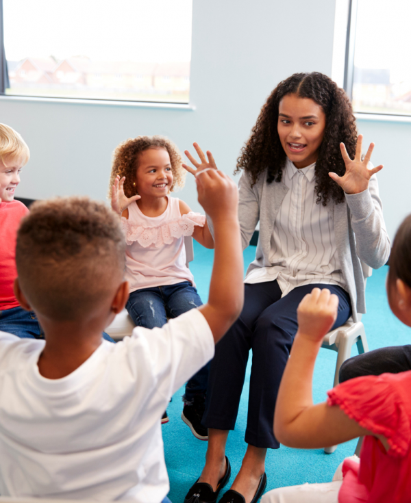 Group of children sit in a circle holding both hands up like stars with psychologist leading activity