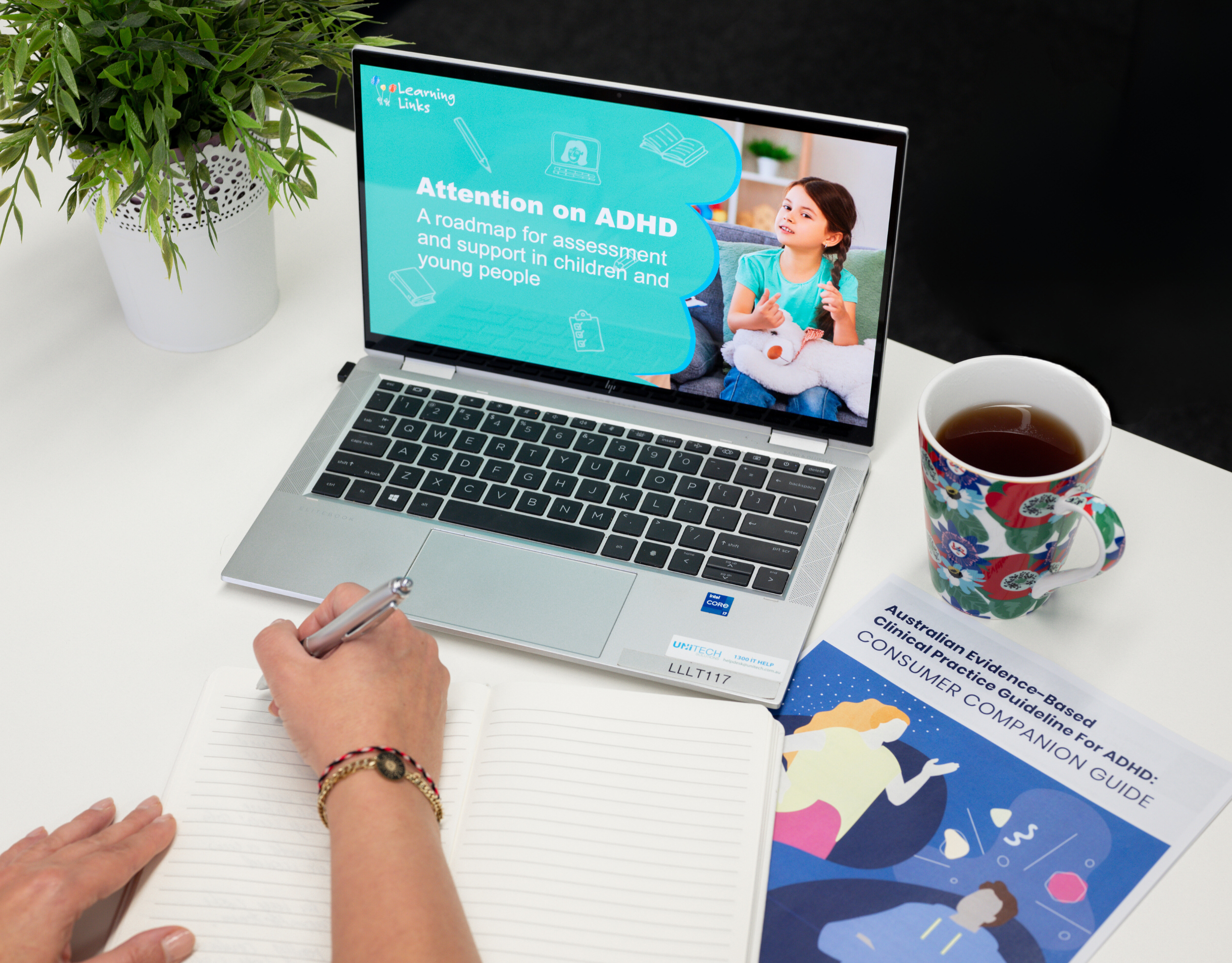 Laptop with course cover slide displayed while person's hand prepares to write notes in a book