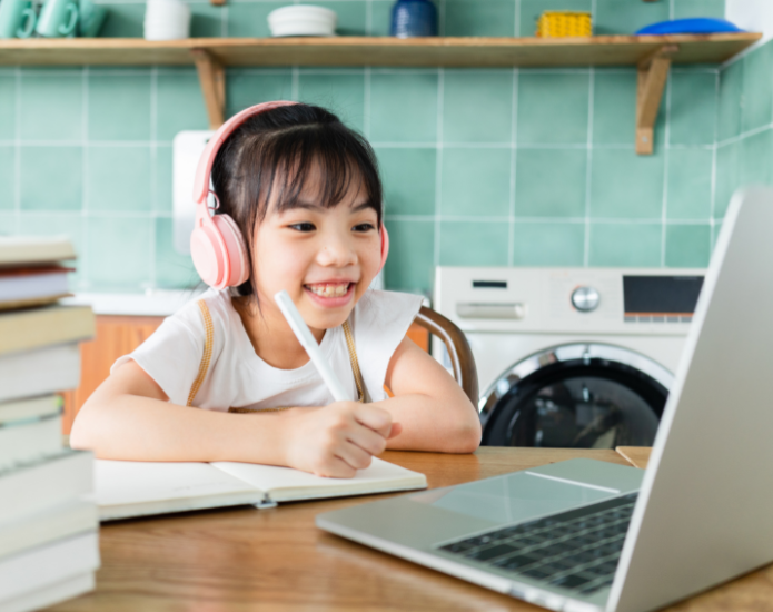 Young girl wearing headphones and looking at laptop screen while preparing to write on a notebook